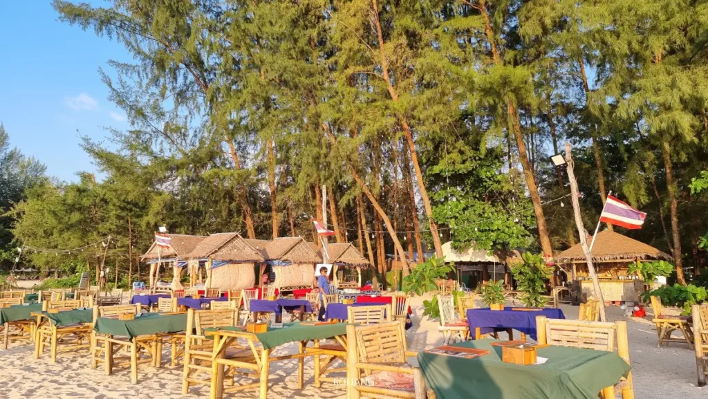 โต๊ะและเก้าอี้ไม้กลางแจ้งพร้อมผ้าคลุมถูกจัดวางบนชายหาดทรายริมทะเลสีคราม อยู่ด้านหน้าของกระท่อมและต้นไม้สูง โดยมีธงชาติไทยหลายผืนปรากฏอยู่ในฉาก.