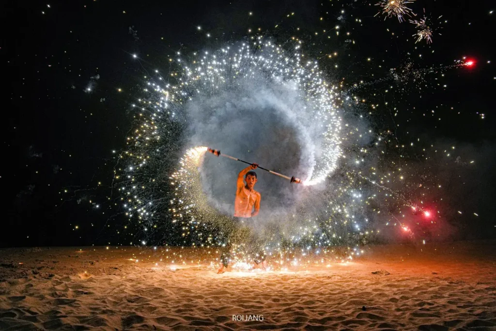 บุคคลหนึ่งแสดงท่าทางด้วยไม้เท้าที่ลุกเป็นไฟบนพื้นทรายในเวลากลางคืน สร้างประกายไฟและควันเป็นวงกลม พร้อมกับมีดอกไม้ไฟปรากฏให้เห็นอยู่เบื้องหลังใกล้ทะเลสีคราม.