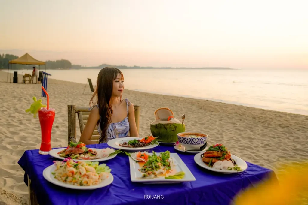 ผู้หญิงคนหนึ่งนั่งอยู่ที่โต๊ะบนชายหาดริมทะเลสีคราม พร้อมจานอาหารทะเล, สลัด, มะพร้าว, และเครื่องดื่มสีแดง กำลังเพลิดเพลินกับมื้ออาหารของเธอในระหว่างพระอาทิตย์ตกดิน.