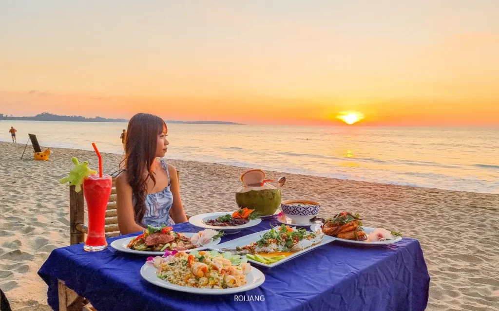 ผู้หญิงคนหนึ่งนั่งอยู่ที่โต๊ะบนชายหาด มีอาหารและเครื่องดื่มหลากหลายชนิดวางอยู่ มองดูพระอาทิตย์ตกดินเหนือทะเลสีคราม.