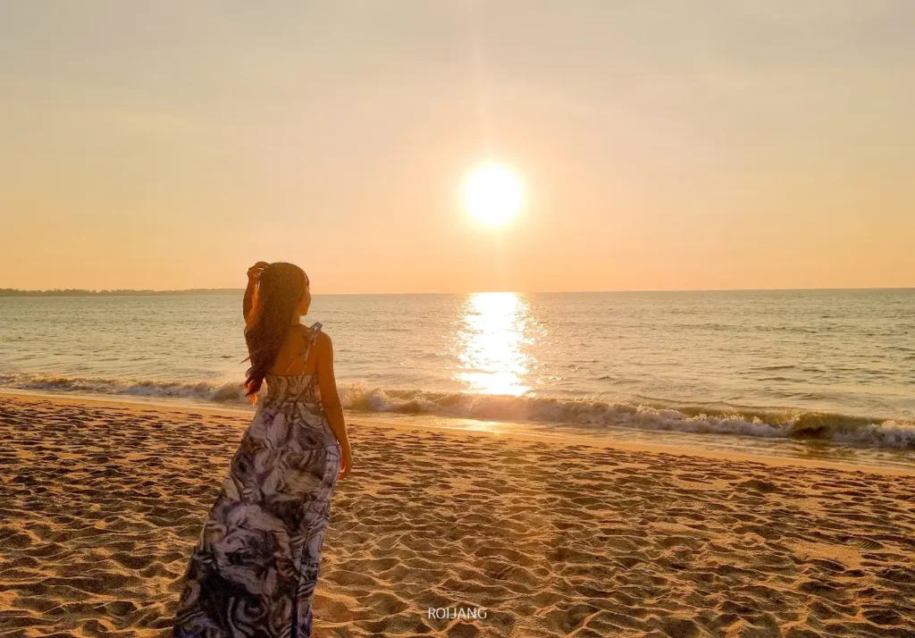 บุคคลในชุดยาว ยืนอยู่บนชายหาดทราย กำลังมองดูพระอาทิตย์ตกดินเหนือทะเลสีคราม.