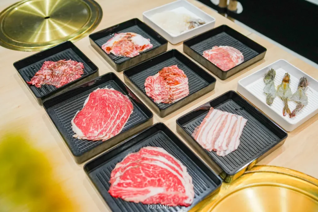 เนื้อสัตว์และอาหารทะเลดิบหลากหลายชนิด รวมถึงเนื้อวัวหั่นบาง หมูสามชั้น และกุ้ง จัดเรียงในถาดสีดำบนโต๊ะที่ร้านบูนต้า กริลล์ พร้อมสำหรับการย่าง.