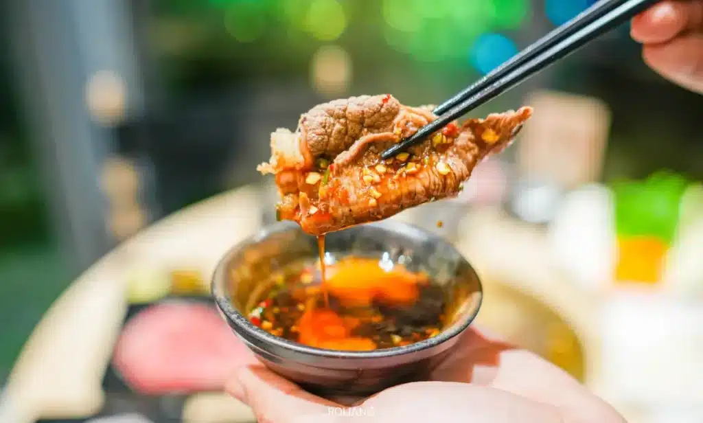 มือใช้ตะเกียบคีบชิ้นเนื้อวัวที่ปรุงสุกและราดซอสจากร้านบูนต้า กริลล์ จุ่มลงในชามเล็กที่เต็มไปด้วยน้ำจิ้ม.