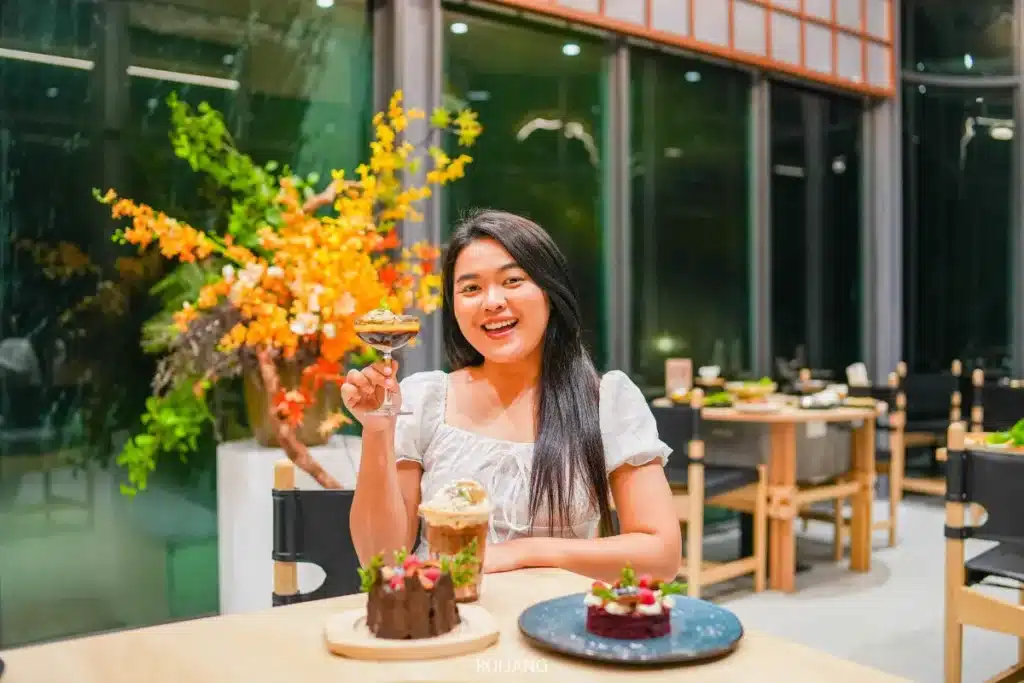 ผู้หญิงคนหนึ่งนั่งอยู่ที่โต๊ะในร้านบูนตา กริลล์ ยกแก้วของหวานขึ้นมายิ้มอยู่ โดยมีของหวานต่าง ๆ อยู่ข้างหน้าเธอ.