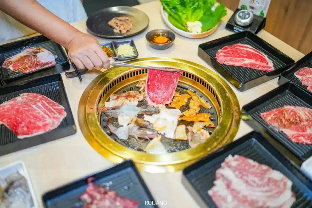 คนกำลังย่างเนื้อสัตว์และอาหารทะเลหลากหลายชนิดบนเตาย่างวงกลม Boonta Grill ล้อมรอบด้วยจานเนื้อวัวสดและเครื่องเคียงรสชาติเข้มข้น.