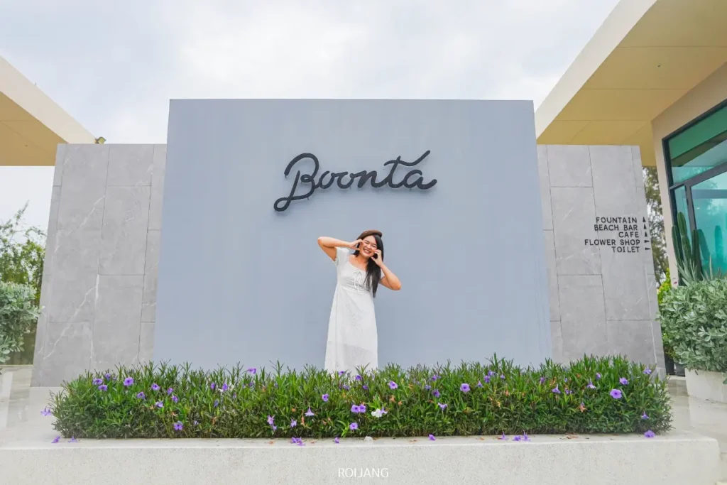 ผู้หญิงในชุดสีขาวโพสท่าเล่นอย่างสนุกสนานหน้าผนังสีเทาที่มีคำว่า "บูนตา" เขียนไว้ ล้อมรอบด้วยดอกไม้สีม่วงและป้ายบอกทางไปยังสิ่งอำนวยความสะดวกต่าง ๆ รวมถึงบูนตา กริลล์.