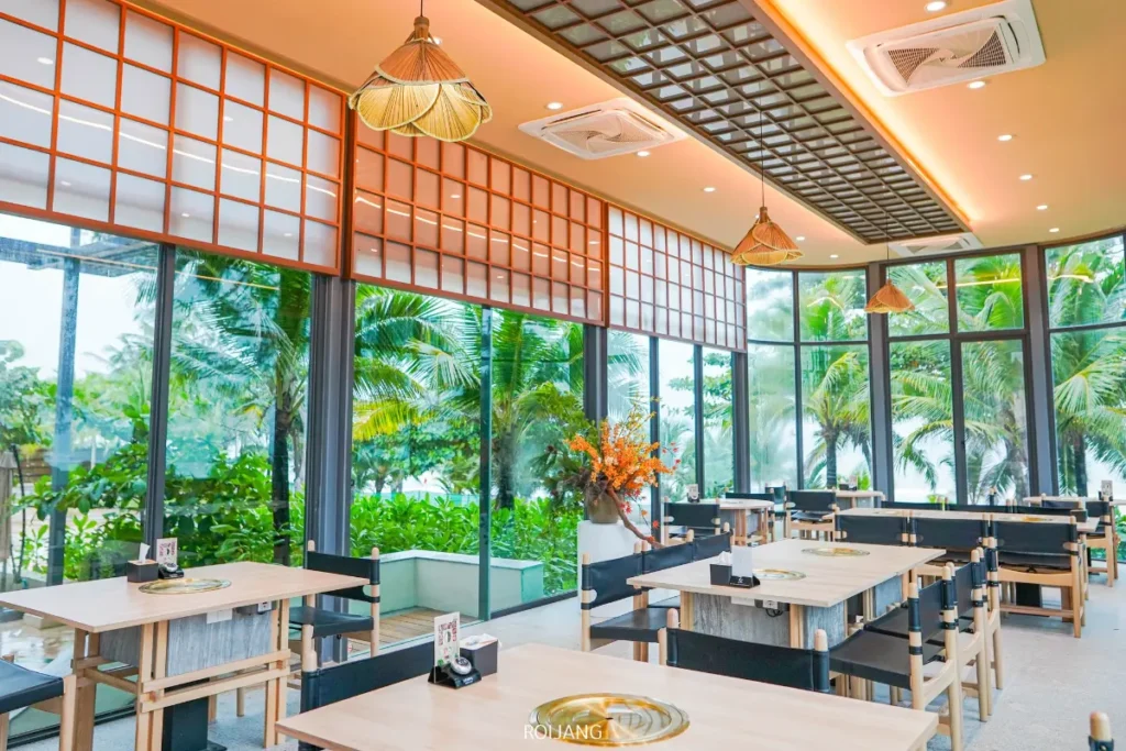 ร้านอาหารสมัยใหม่ภายใน Boonta Grill พร้อมโต๊ะและเก้าอี้ไม้ หน้าต่างบานใหญ่ที่มองเห็นต้นไม้และต้นปาล์ม พร้อมไฟเพดานและของตกแต่งในโทนอบอุ่น.