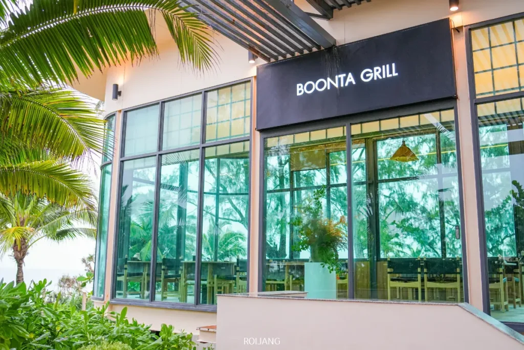 ภายนอกของร้าน Boonta Grill โดดเด่นด้วยกระจกบานใหญ่ล้อมรอบด้วยพืชพรรณเขียวชอุ่มและต้นปาล์ม สะท้อนบรรยากาศเขตร้อนที่เชิญชวนของร้านอาหารได้อย่างสมบูรณ์แบบ.