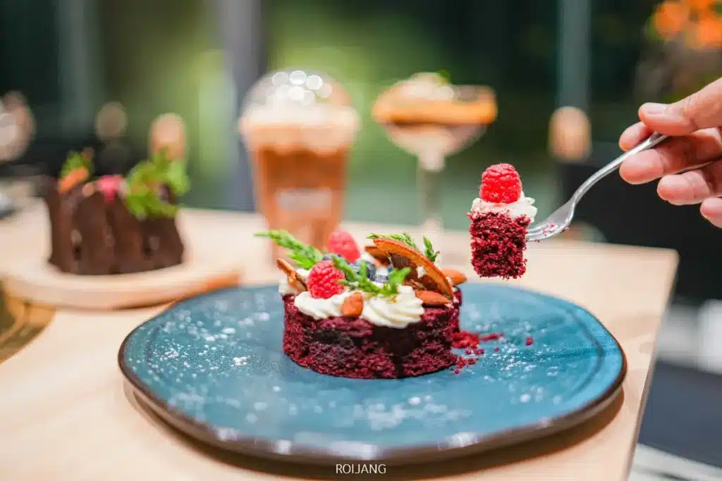 มือหนึ่งถือส้อมที่มีเค้กเรดเวลเวทคำหนึ่งซึ่งมีครีมและราสป์เบอร์รี่จากร้าน Boonta Grill อยู่ด้านบน ส่วนที่เหลือของเค้กวางอยู่บนจานสีน้ำเงิน เครื่องดื่มและของหวานอีกอย่างหนึ่งอยู่ในฉากหลัง.