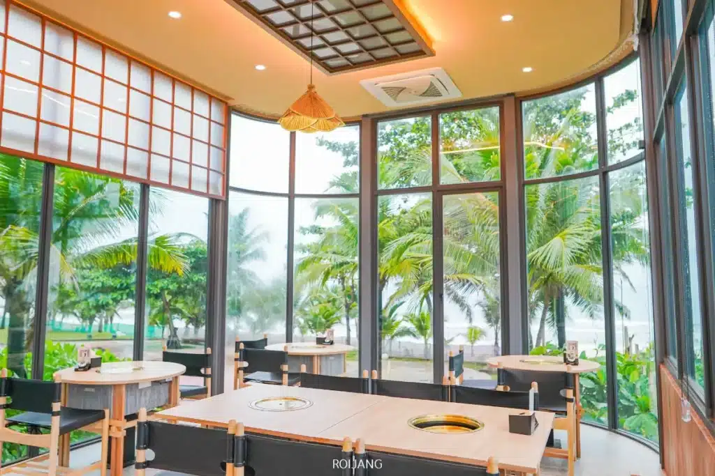 ภายในร้านอาหารที่สว่างและทันสมัย พร้อมหน้าต่างบานใหญ่จากพื้นจรดเพดานที่ Boonta Grill มีโต๊ะกลมและต้นปาล์มที่มองเห็นได้จากภายนอก.