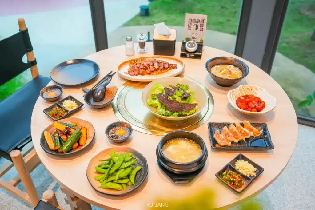 โต๊ะกลมที่ Boonta Grill ถูกจัดเตรียมไว้พร้อมอาหารเกาหลีหลากหลายชนิด รวมถึงเนื้อสัตว์ย่าง ซุป เกี๊ยว ผัก น้ำจิ้ม และเครื่องเคียงต่างๆ ในพื้นที่รับประทานอาหารที่สว่างไสวด้วยหน้าต่างบานใหญ่.