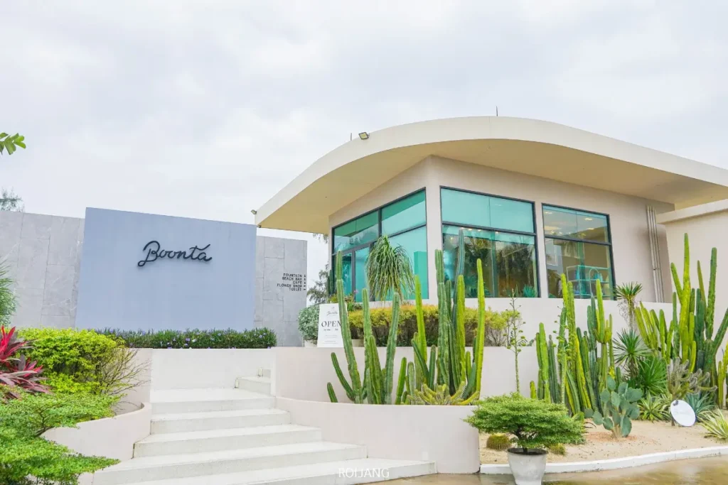 อาคารสมัยใหม่ที่มีหน้าต่างบานใหญ่และหลังคาโค้งล้อมรอบด้วยกระบองเพชรและพืชสีเขียว; มีป้ายใกล้ทางเข้าเขียนว่า "บูนตา กริลล์.