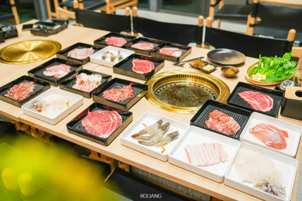 เนื้อสัตว์ดิบหลากหลายชนิด อาหารทะเล และผักที่จัดเรียงในถาดรอบเตาย่างบนโต๊ะที่ร้านบูนต้า กริลล์ พร้อมสำหรับประสบการณ์บาร์บีคิวเกาหลีแท้ๆ.