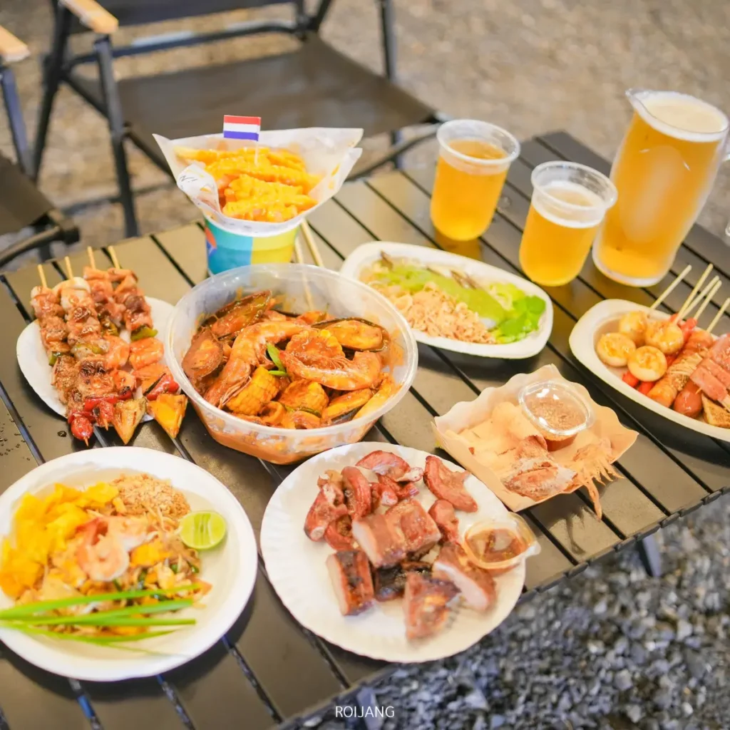 โต๊ะที่มีเนื้อสัตว์ย่างหลากหลายชนิดเสียบไม้ กุ้งทอด ข้าว ไส้กรอกหั่นเป็นชิ้น น้ำจิ้ม และเบียร์สามแก้วพร้อมเหยือกหนึ่ง—เหมาะสำหรับการรวมตัวกับเพื่อนและแบ่งปันช่วงเวลาแห่งความสุข.