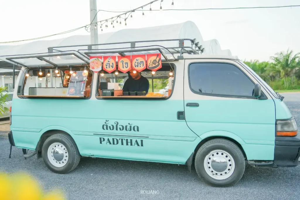 รถขายอาหารสีฟ้าอมเขียวที่มีคำว่า "PADTHAI" และ "มีสุข" เขียนอยู่ด้านข้างจอดอยู่กลางแจ้ง โดยมีพ่อค้าแม่ค้าเตรียมอาหารอยู่ภายใน.
