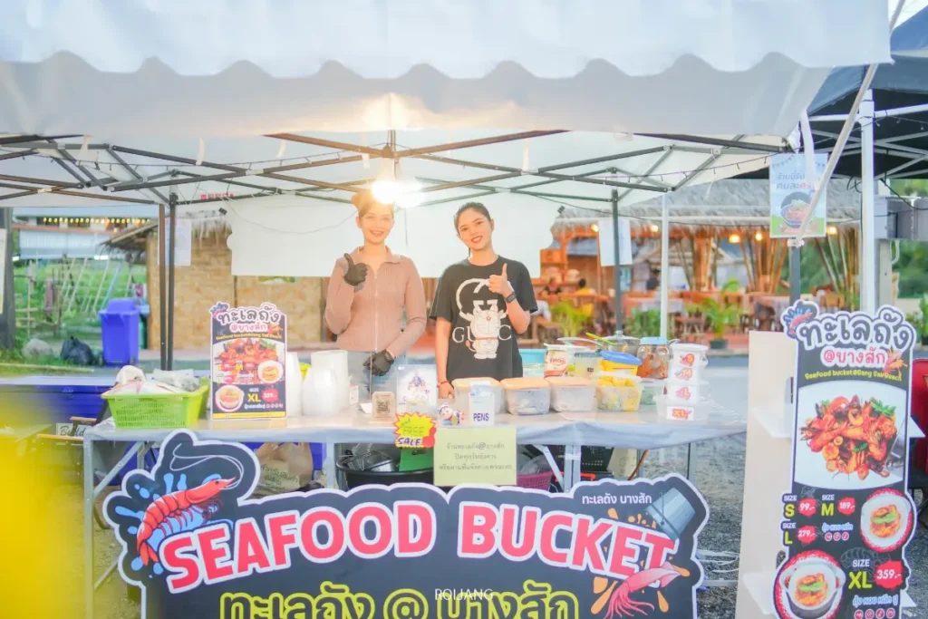 พ่อค้าแม่ค้าสองคนยืนอยู่หลังร้านขายอาหารทะเลในตะกร้าที่ตลาดกลางแจ้ง ยิ้มอย่างมีความสุขและโพสท่าถ่ายรูป โดยมีภาชนะใส่อาหารและป้ายเมนูต่างๆ อยู่บนโต๊ะ.