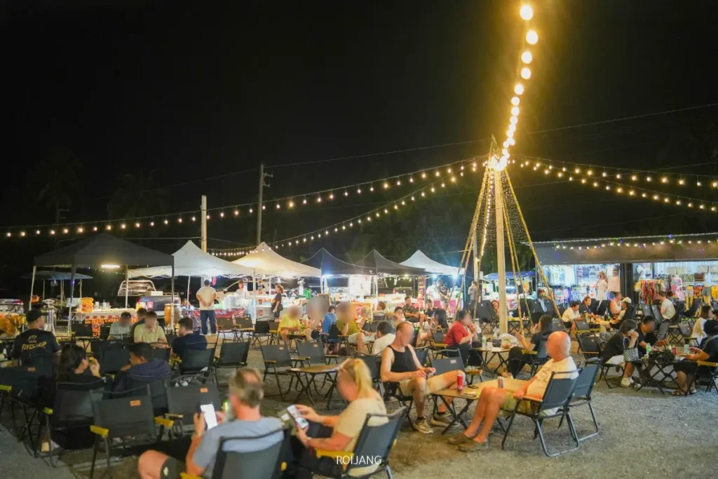 ผู้คนนั่งอยู่ที่โต๊ะใต้ไฟประดับสายในตลาดกลางคืนกลางแจ้ง เพลิดเพลินกับอาหารอร่อยจากแผงขายและเต็นท์ในฉากหลัง สร้างบรรยากาศที่คึกคักเต็มไปด้วยความสุข.