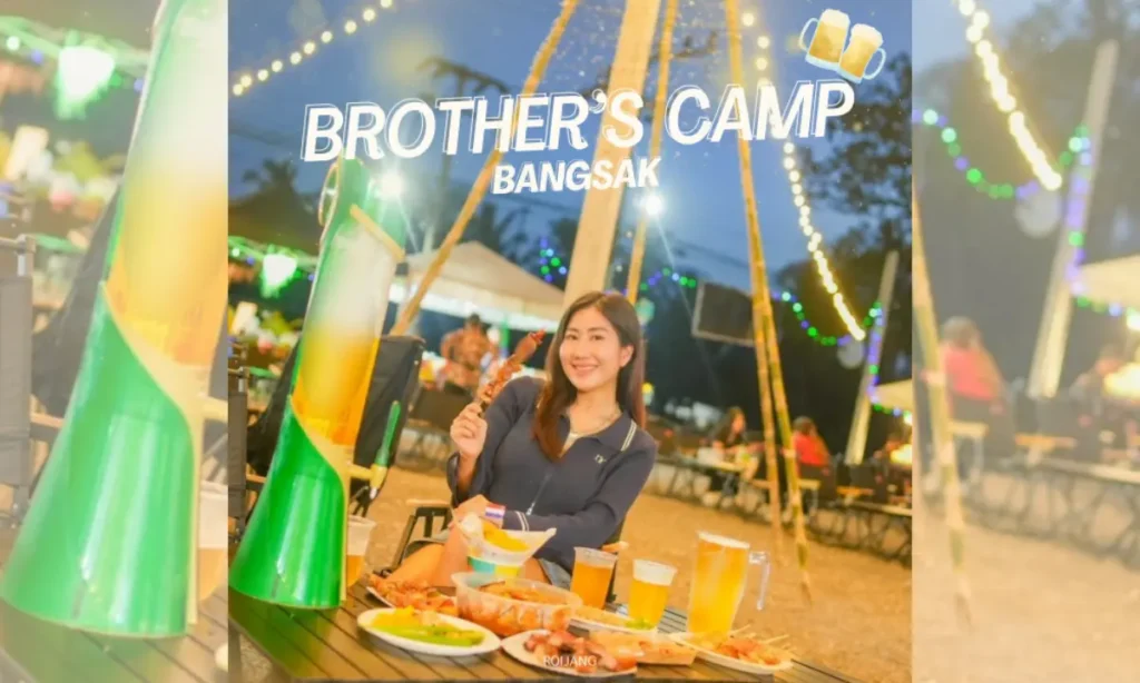 ผู้หญิงคนหนึ่งนั่งอยู่ที่โต๊ะกลางแจ้งพร้อมอาหารและเครื่องดื่มในยามค่ำคืน ยิ้มอย่างมีความสุขให้กล้อง "Brother's Camp Bangsak" เขียนไว้ด้านบนพร้อมกับภาพแก้วเบียร์.
