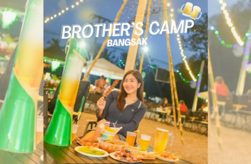 ผู้หญิงคนหนึ่งนั่งอยู่ที่โต๊ะกลางแจ้งพร้อมอาหารและเครื่องดื่มในยามค่ำคืน ยิ้มอย่างมีความสุขให้กล้อง "Brother's Camp Bangsak" เขียนไว้ด้านบนพร้อมกับภาพแก้วเบียร์.