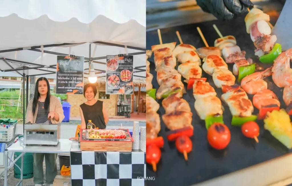 ผู้หญิงสองคนยืนอยู่หลังแผงขายอาหารริมทาง โดยมีไม้เสียบอาหารวางโชว์อยู่ บรรยากาศเต็มไปด้วยความสุข; ภาพระยะใกล้ของไม้เสียบเนื้อย่างและผักที่วางอยู่ทางขวา ส่องประกายแวววาว.