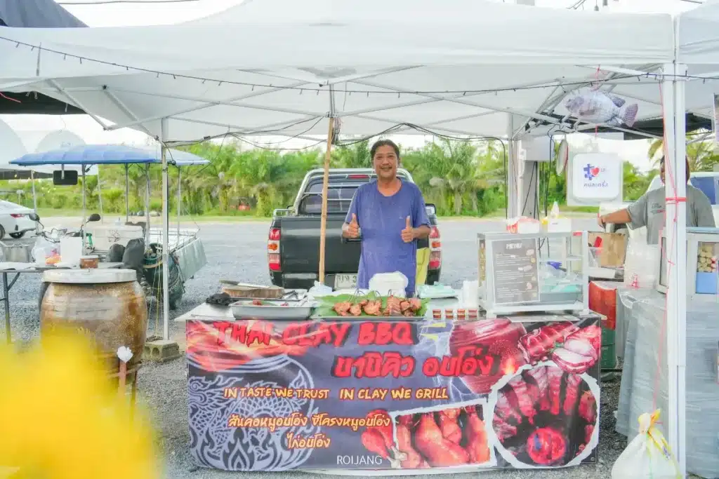 คนยืนอยู่หลังแผงขายอาหารบาร์บีคิวดินเผาไทย มีเตาย่างและของต่างๆ บนเคาน์เตอร์ พร้อมทำท่าชูนิ้วโป้งแบบ "มีสุข" ใต้เต็นท์สีขาว.