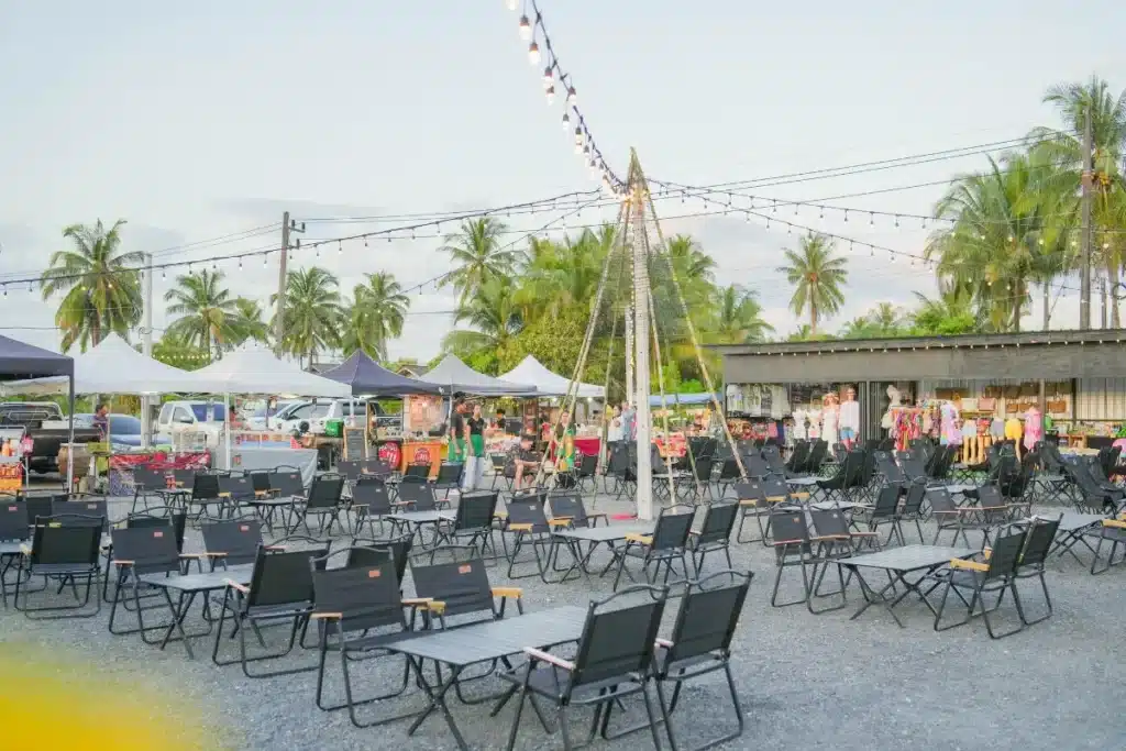 พื้นที่นั่งกลางแจ้งที่มีแถวของเก้าอี้และโต๊ะสีดำ, ไฟประดับแขวนอยู่เหนือศีรษะ, ร้านค้าตลาดและต้นปาล์มเป็นฉากหลัง, สร้างบรรยากาศที่สุขสบายเหมาะสำหรับการผ่อนคลายกับเพื่อน ๆ.