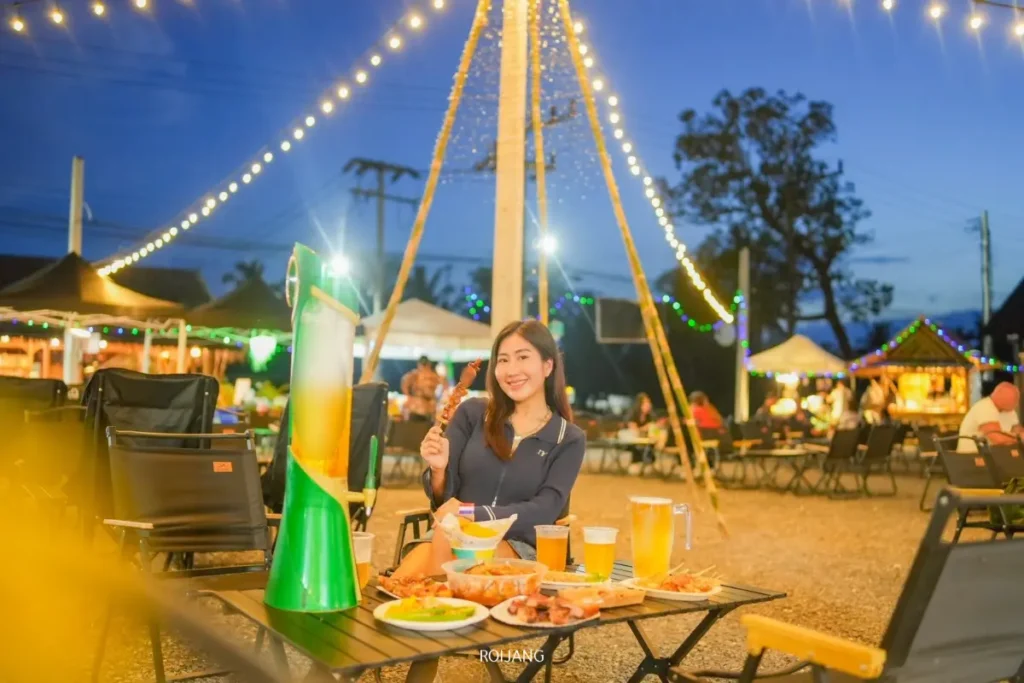 ผู้หญิงคนหนึ่งนั่งอยู่ที่โต๊ะกลางแจ้งพร้อมอาหารและเครื่องดื่มในยามค่ำคืน ล้อมรอบด้วยไฟประดับและโต๊ะอื่น ๆ ในบรรยากาศที่เต็มไปด้วยความสุขและเทศกาล.