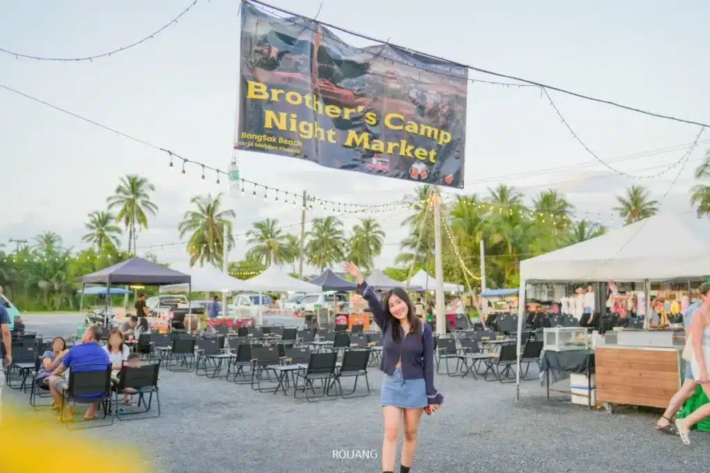 ผู้หญิงคนหนึ่งยืนและโบกมืออยู่ใต้ป้ายขนาดใหญ่ที่เขียนว่า "ตลาดกลางคืนค่ายบราเธอร์" ที่ตลาดกลางแจ้ง บรรยากาศเต็มไปด้วยความสุข มีเต็นท์ เก้าอี้ และต้นปาล์มเป็นฉากหลัง.