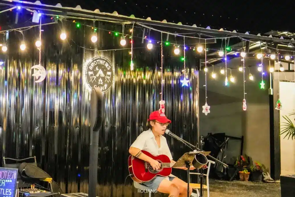 บุคคลหนึ่งสวมหมวกสีแดง ดูมีความสุข กำลังเล่นกีตาร์โปร่งและร้องเพลงผ่านไมโครโฟน นั่งอยู่หน้าผนังสีดำที่ประดับด้วยไฟประดับและเครื่องตกแต่งในยามค่ำคืน.