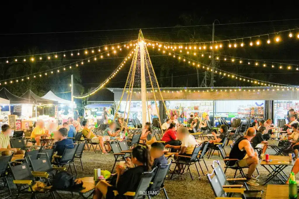 ตลาดนัดกลางคืนกลางแจ้งที่มีผู้คนนั่งที่โต๊ะใต้แสงไฟประดับ ล้อมรอบด้วยแผงขายอาหารและบูธตลาด สร้างบรรยากาศครึกครื้นและมีความสุขสำหรับทุกคนได้เพลิดเพลิน.