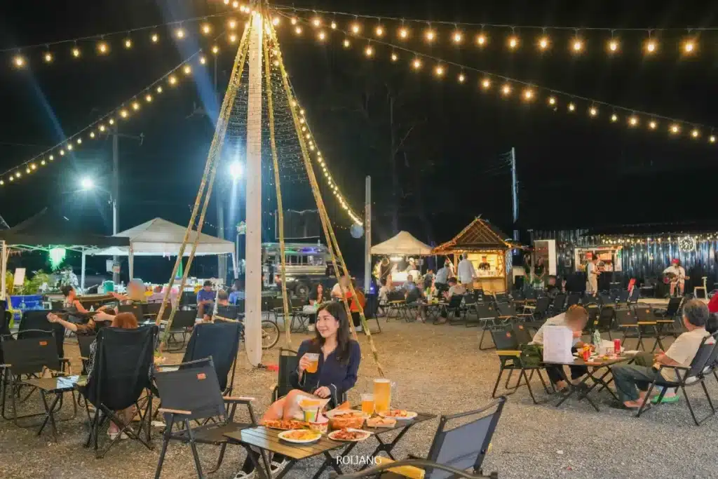 ผู้คนกำลังรับประทานอาหารกลางแจ้งในยามค่ำคืนภายใต้แสงไฟประดับสาย นั่งที่โต๊ะพร้อมอาหารและเครื่องดื่ม ทุกคนกำลังเพลิดเพลินกับบรรยากาศที่สุขสันต์; ร้านอาหารและเต็นท์สามารถมองเห็นได้ในพื้นหลัง.