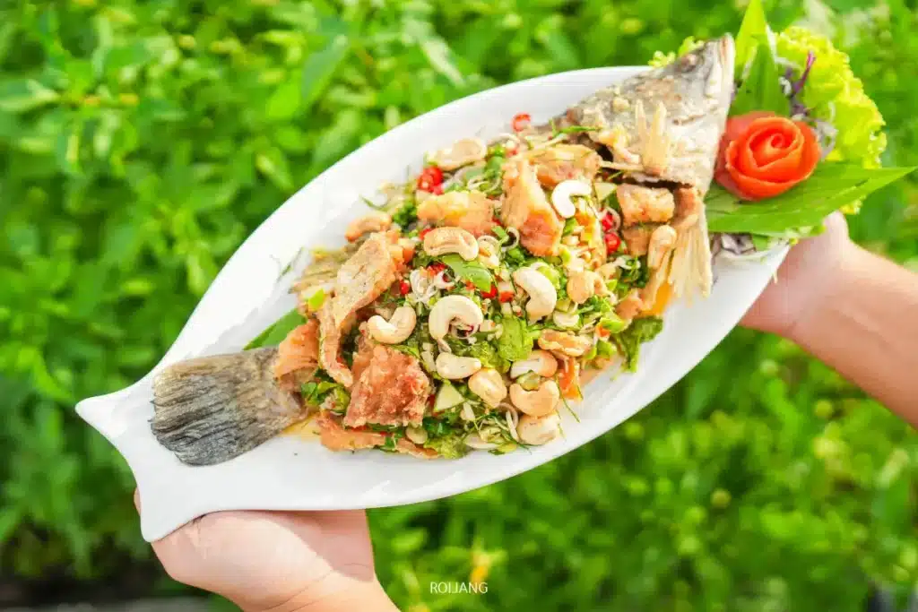 บุคคลหนึ่งถือจานสีขาวที่มีปลาทอดทั้งตัววางอยู่ด้านบน โรยด้วยสมุนไพร เม็ดมะม่วงหิมพานต์ และพริกหั่นบางๆ ตกแต่งด้วยกุหลาบที่ทำจากมะเขือเทศ—สดใหม่จากตลาดแคมป์ของพี่ชาย.