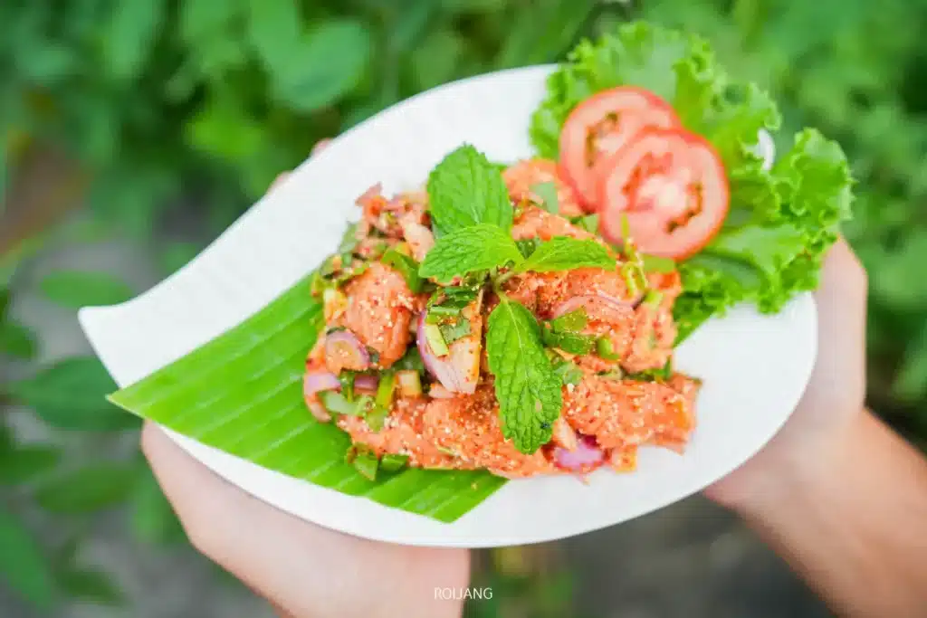 จานสลัดเนื้อสับเผ็ดจากตลาดแคมป์บราเธอร์ ตกแต่งด้วยใบสะระแหน่ หอมหัวใหญ่หั่นบาง ผักกาดหอม และมะเขือเทศ เสิร์ฟบนใบตอง.