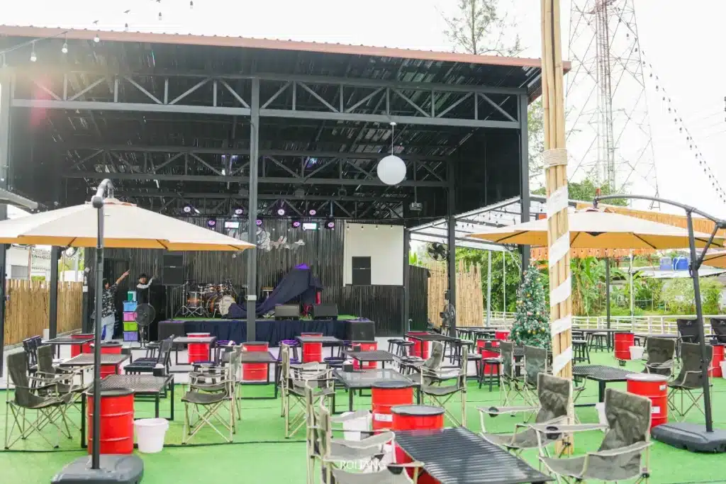 สถานที่จัดงานกลางแจ้งที่ Brother's Camp Market พร้อมเวทีที่มีหลังคา เครื่องดนตรี และพื้นที่นั่งที่มีโต๊ะ เก้าอี้ และร่มบนสนามหญ้าเทียม ไฟเวทีเปิดสว่าง รายล้อมด้วยต้นไม้ในฉากหลัง.