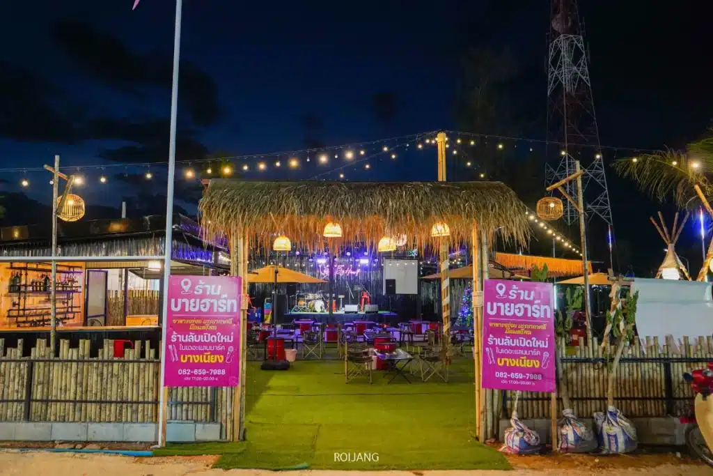 ร้านอาหารกลางแจ้ง Brother's Camp Market มีทางเข้าเป็นหลังคามุงจาก ตกแต่งด้วยไฟประดับ และป้ายภาษาไทย ถ่ายภาพในเวลากลางคืนขณะมีลูกค้านั่งรับประทานอาหารอยู่ภายใน.