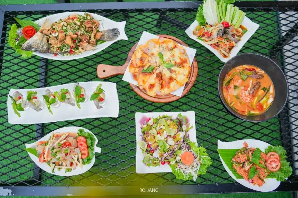 อาหารไทยหลากหลายชนิดและพิซซ่าจาก Brother's Camp Market จัดเรียงอยู่บนโต๊ะเหล็กสีดำ ประกอบด้วยสลัด ซุป ปลาเผา และอาหารเรียกน้ำย่อยที่ตกแต่งด้วยสมุนไพรสดและผัก.