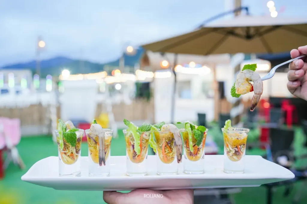 ถาดอาหารเรียกน้ำย่อยกุ้งในแก้วช็อตตกแต่งด้วยสมุนไพรถูกวางไว้กลางแจ้งที่ตลาดแคมป์ของบราเธอร์ โดยมีแสงไฟพร่ามัวและร่มเป็นฉากหลัง.