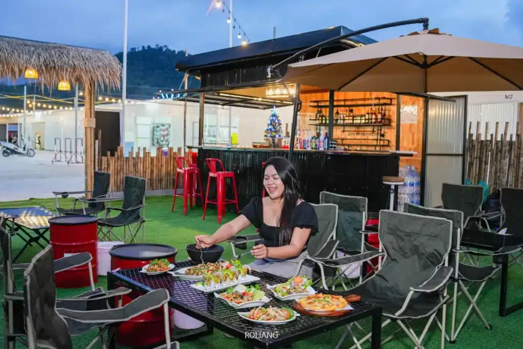 ผู้หญิงคนหนึ่งนั่งอยู่ที่โต๊ะกลางแจ้งซึ่งจัดเตรียมอาหารหลากหลายชนิดไว้ รวมถึงสลัดและพิซซ่า ใกล้กับเคาน์เตอร์บาร์สีดำและร่มสนามบนหญ้าเทียมที่ตลาดแคมป์ของบราเธอร์.