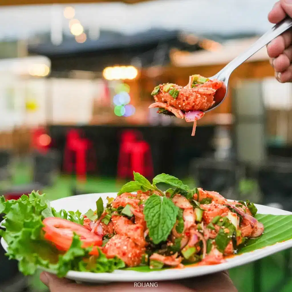 มือหนึ่งถือช้อนที่ตักสลัดเนื้อสับรสเผ็ดไว้เต็มช้อน เหนือจานที่ตกแต่งด้วยผักกาดหอม ใบสะระแหน่ และมะเขือเทศ ณ ตลาดแคมป์ของพี่ชาย ท่ามกลางบรรยากาศกลางแจ้งที่ชวนให้น่าเข้าไปสัมผัส.