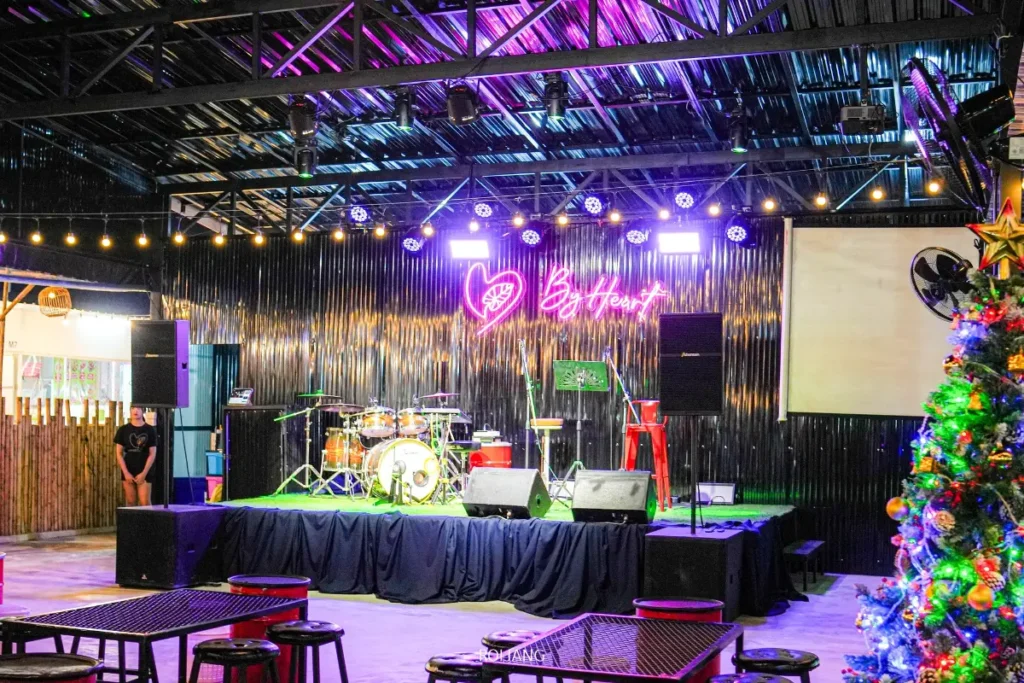 เวทีที่จัดเตรียมไว้พร้อมกลอง ไมโครโฟน และลำโพง ภายใต้แสงไฟสีสันสดใสที่ Brother's Camp Market โดยมีป้ายไฟนีออน "By Heart" อยู่บนผนังด้านหลัง และต้นคริสต์มาสที่ตกแต่งอย่างสวยงามอยู่ด้านหน้า.