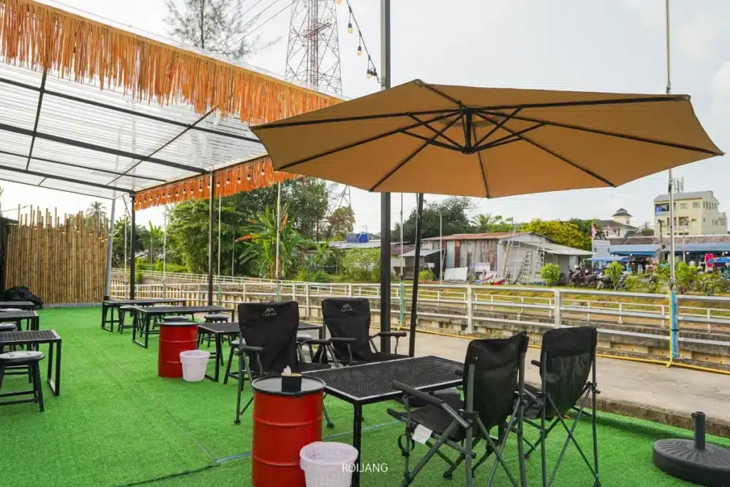 พื้นที่นั่งกลางแจ้งที่ Brother's Camp Market พร้อมโต๊ะและเก้าอี้สีดำ ร่มขนาดใหญ่สีเบจ เก้าอี้บาร์ทรงถังสีแดง และพื้นหญ้าเทียมใกล้รั้วและถนน.