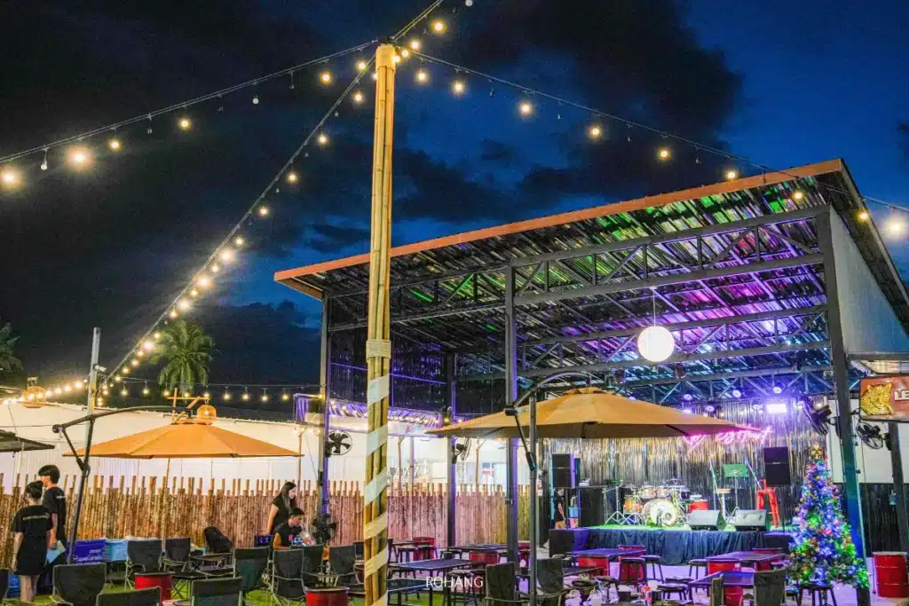 เวทีกลางแจ้งที่ตลาดแคมป์บราเธอร์ พร้อมเครื่องดนตรีที่จัดวางไว้ใต้หลังคาเหล็ก มีไฟประดับแขวนอยู่เหนือศีรษะ โต๊ะและเก้าอี้จัดเรียงไว้เรียบร้อย ผู้คนกำลังเตรียมสถานที่ในช่วงพลบค่ำ.