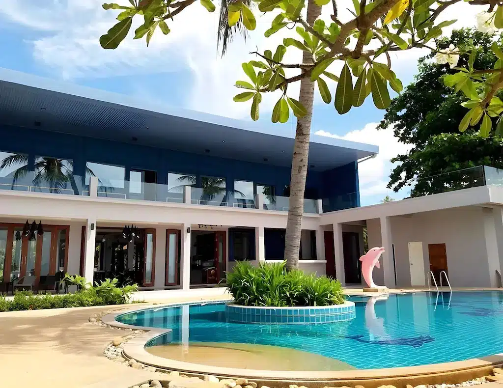บ้านสองชั้นสมัยใหม่พร้อมหน้าต่างบานใหญ่ สระว่ายน้ำ ต้นปาล์ม และประติมากรรมปลาโลมาสีชมพูในสระว่ายน้ำ—เหมาะสำหรับผู้ที่มองหา รีสอร์ทขนอม หรือ ที่พักขนอม ที่มีสไตล์ภายใต้ท้องฟ้าที่มีเมฆบางส่วน.