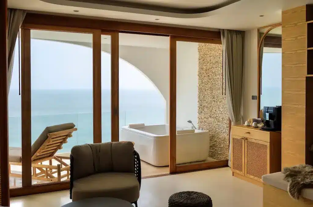 ห้องพักโรงแรมสมัยใหม่ที่ โรงแรมขนอม พร้อมการตกแต่งภายในสไตล์ทันสมัย เก้าอี้เลานจ์บนระเบียง อ่างอาบน้ำติดหน้าต่างบานใหญ่ และวิวทะเลที่สวยงามเป็นฉากหลัง.