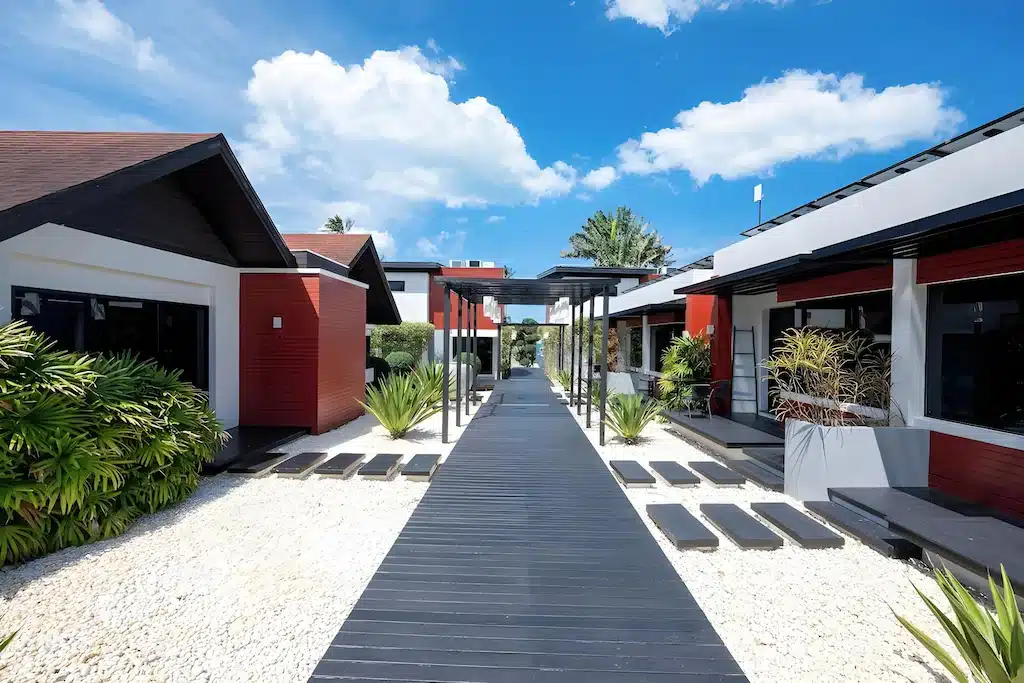 ทางเดินวิลล่าสมัยใหม่พร้อมทางเดินไม้สีดำ กรวดสีขาว พืชสีเขียว และอาคารสีแดงและขาวใต้ท้องฟ้าสีฟ้าสดใส—เหมาะสำหรับการพักผ่อนที่ โรงแรมขนอม หรือผู้ที่มองหา ที่พักในขนอม.