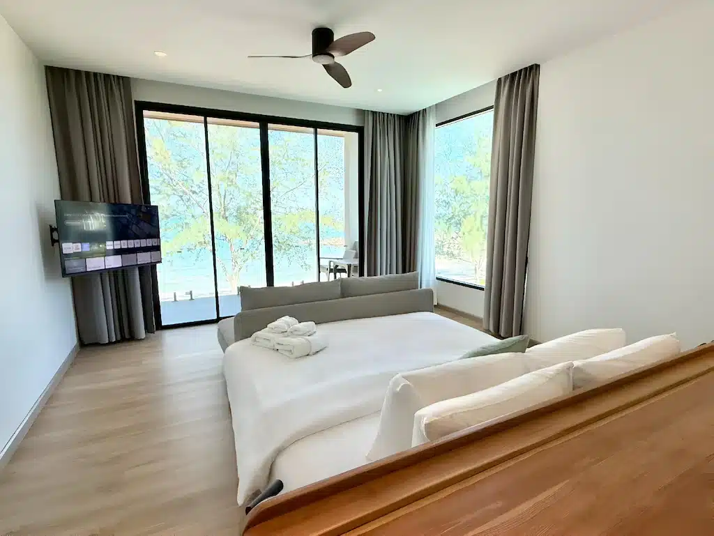 ห้องนอนสมัยใหม่พร้อมเตียงขนาดใหญ่, ทีวี, พัดลมเพดาน, และหน้าต่างบานใหญ่จากพื้นจรดเพดานที่สามารถมองเห็นระเบียงและต้นไม้ภายนอกได้ ผ้าเช็ดตัวถูกพับไว้อย่างเรียบร้อยบนเตียง—เหมาะสำหรับการเข้าพักของคุณที่ ขนอมที่พัก หรือ โรงแรมขนอม.