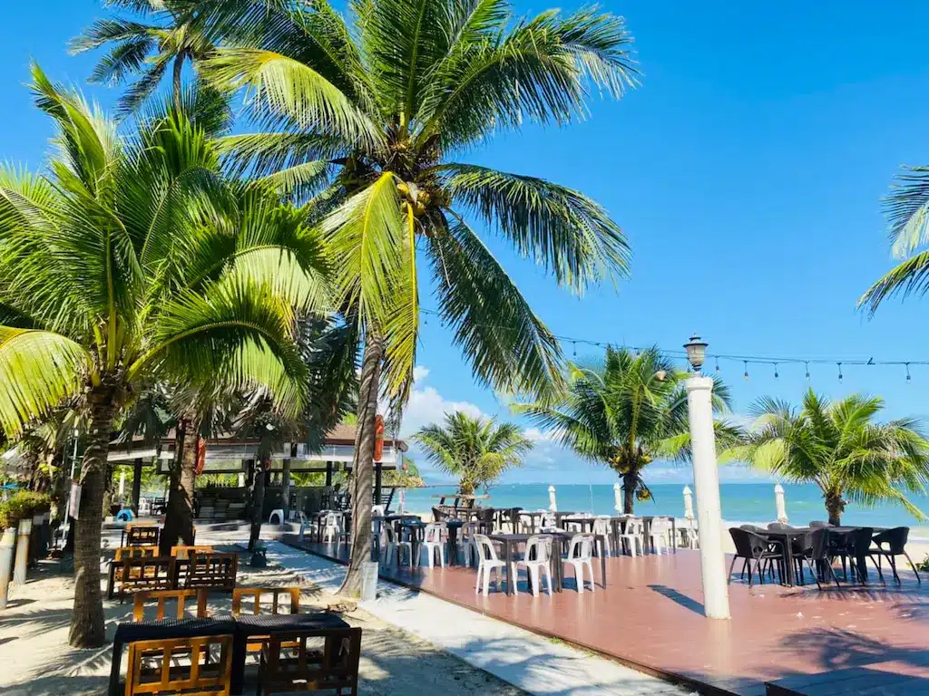 พื้นที่รับประทานอาหารกลางแจ้งริมชายหาดที่รีสอร์ทขนอม พร้อมโต๊ะและเก้าอี้ไม้และพลาสติก ล้อมรอบด้วยต้นปาล์ม มองเห็นท้องฟ้าสีฟ้าสดใสและมหาสมุทร.