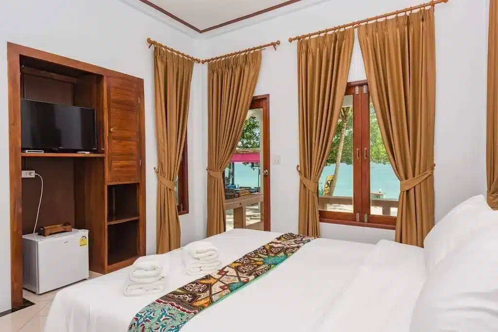 เตียงที่จัดไว้อย่างเรียบร้อยพร้อมผ้าเช็ดตัวพับไว้, ผ้าปูโต๊ะลายสวย, โทรทัศน์, ตู้เย็นขนาดเล็ก, ชั้นวางของไม้, และหน้าต่างบานใหญ่พร้อมผ้าม่านสีน้ำตาลที่มองเห็นน้ำและต้นไม้เขียวขจี—เหมาะสำหรับการพักผ่อนอย่างผ่อนคลายที่รีสอร์ทขนอม.