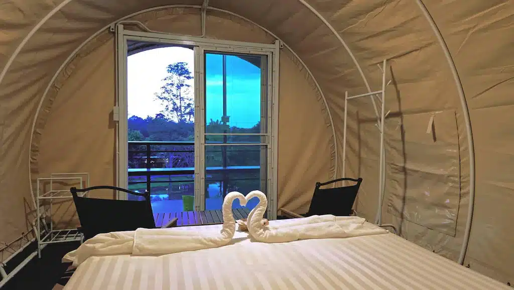 ห้องพักเต็นท์แสนอบอุ่นที่รีสอร์ทเพชรบูรณ์ พร้อมเตียงคู่ ผ้าขนหนูรูปหงส์สองผืนเรียงเป็นรูปหัวใจ ชั้นวางของโลหะ และประตูเปิดโล่งสู่ระเบียงที่มองเห็นต้นไม้และท้องฟ้าสีคราม—เหมาะอย่างยิ่งสำหรับการพักผ่อนที่เพชรบูรณ์ของคุณ.