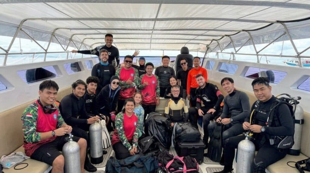 กลุ่มคนในชุดดำน้ำและอุปกรณ์ดำน้ำโพสท่าถ่ายรูปบนเรือ โดยมีถังอากาศดำน้ำและอุปกรณ์ต่างๆ อยู่รอบๆ.